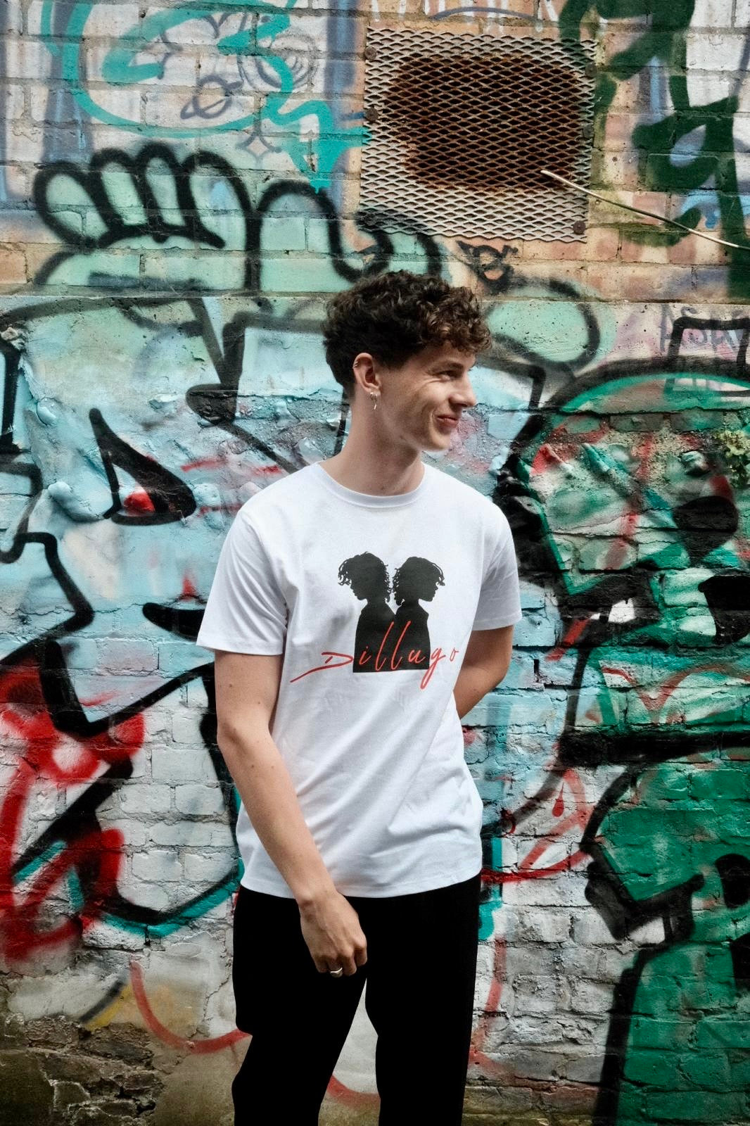 Male model looking left wearing white t-shirt with "Dillugo" written across it (in red) and the signature Dillugo silhouette, on a graffiti backdrop.