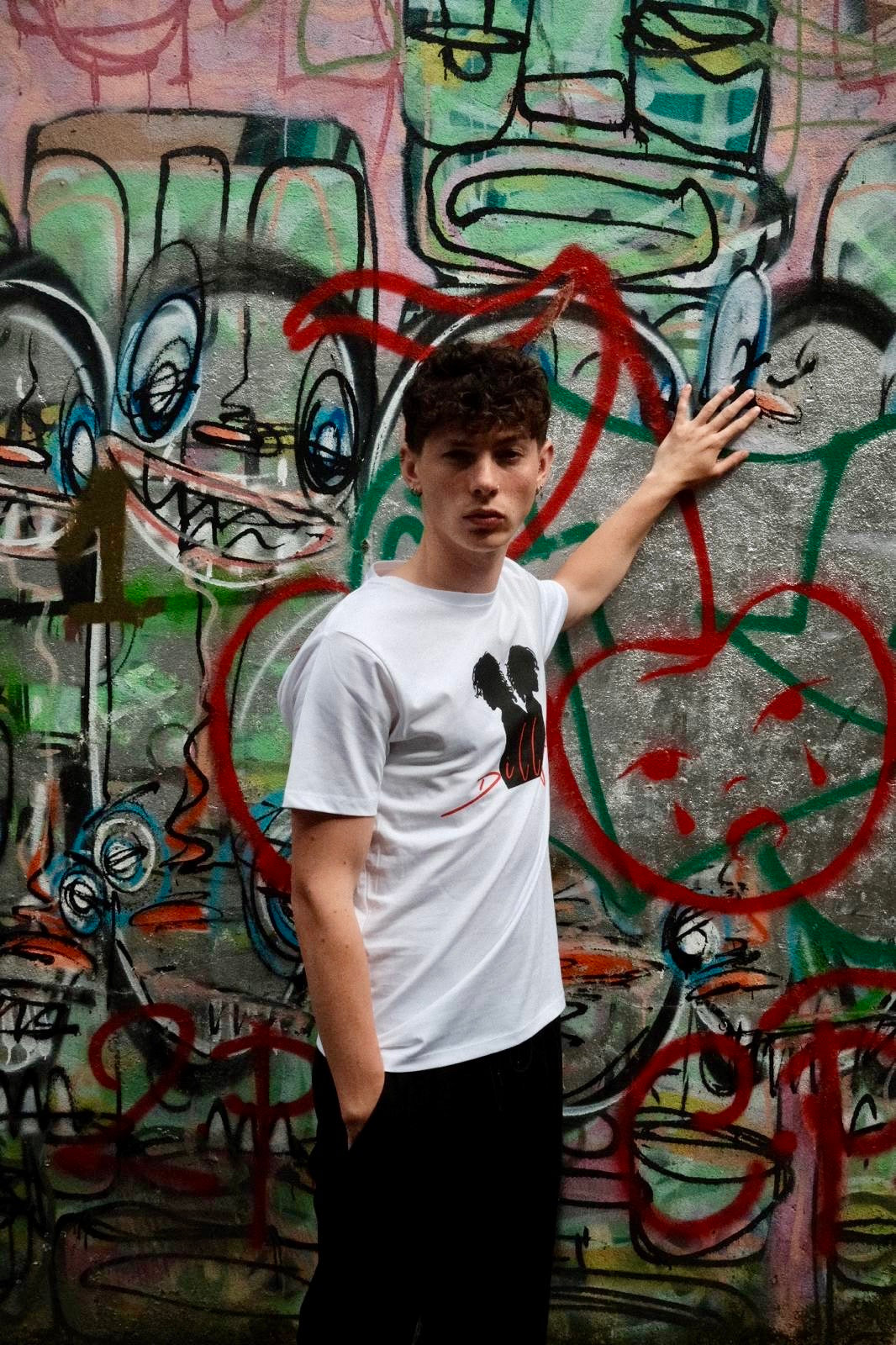 Male model with left hand on wall, wearing white t-shirt with "Dillugo" written across it (in red) and the signature Dillugo silhouette, on a graffiti backdrop.