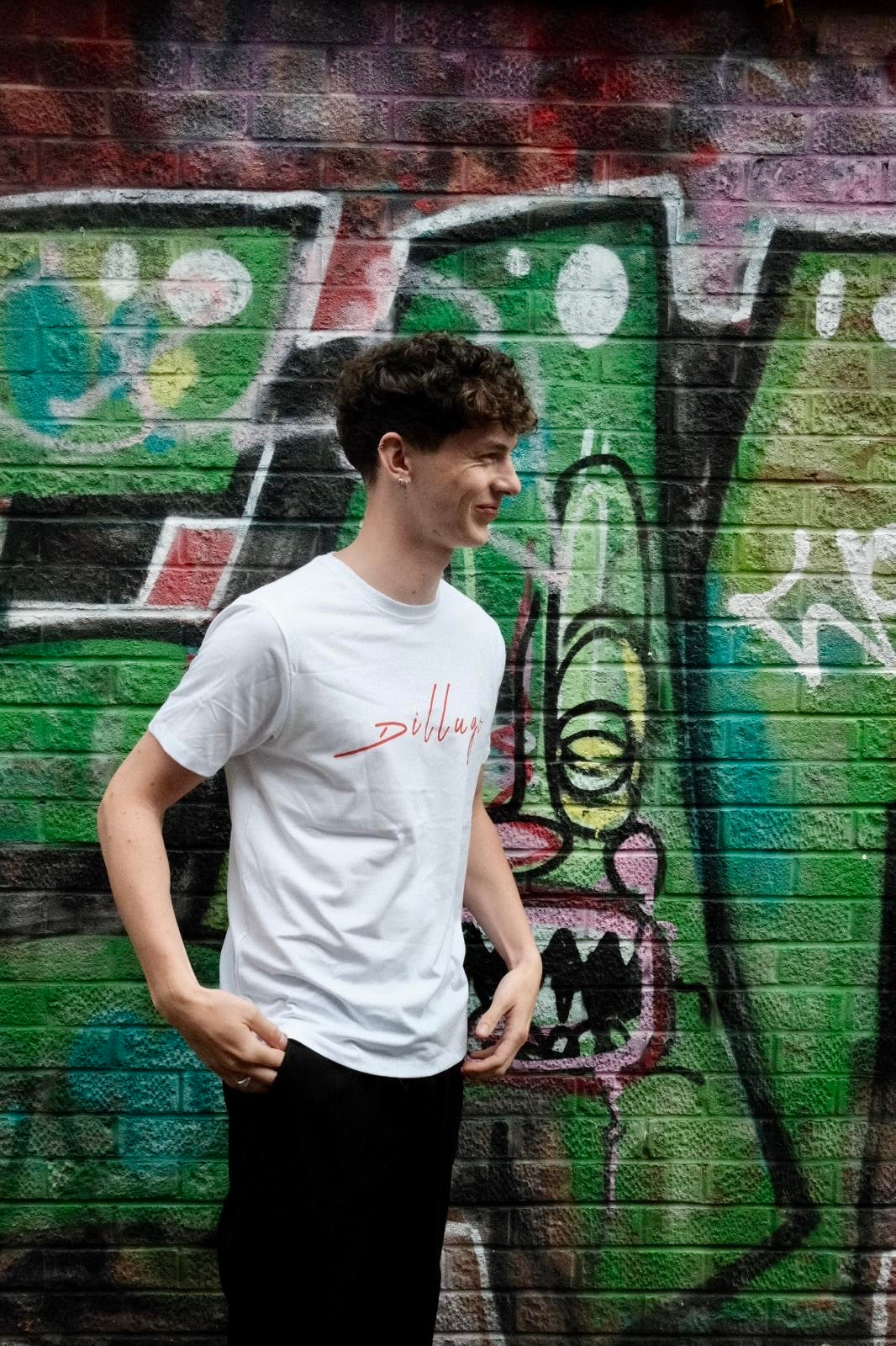 Male model facing his body left wearing white t-shirt with "Dillugo" written across it (in red) on  a graffiti backdrop.