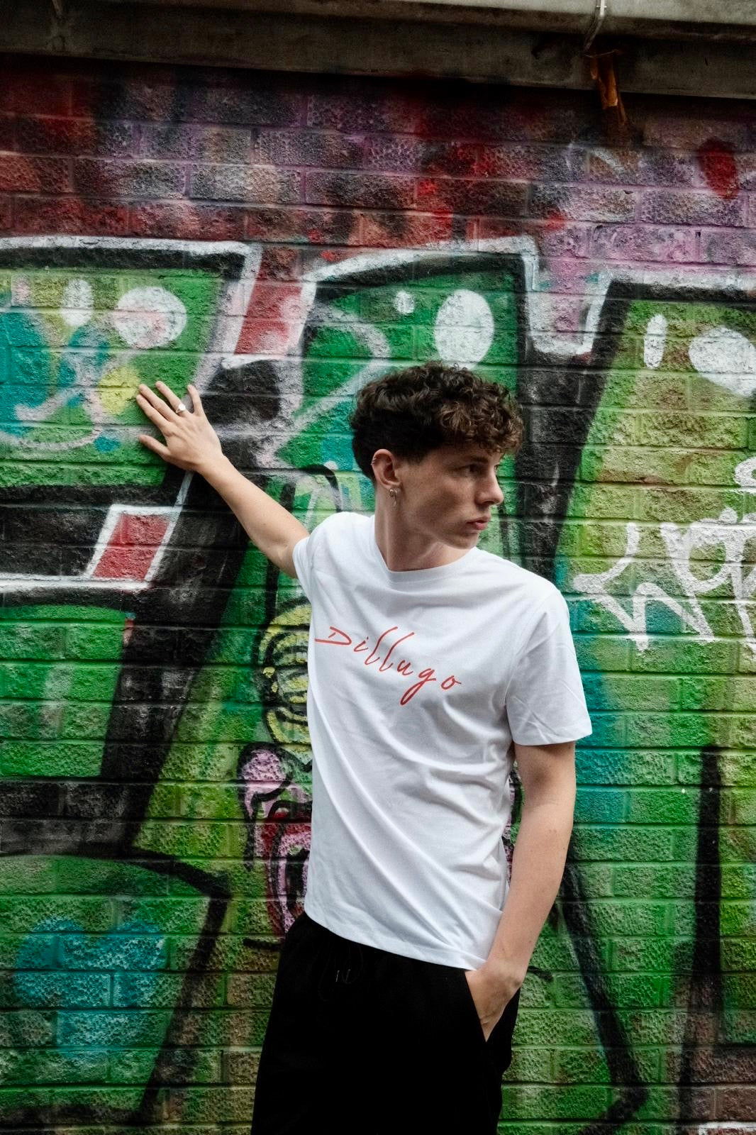 Male model with right hand on wall looking  left wearing white t-shirt with "Dillugo" written across it (in red) on a graffiti backdrop.