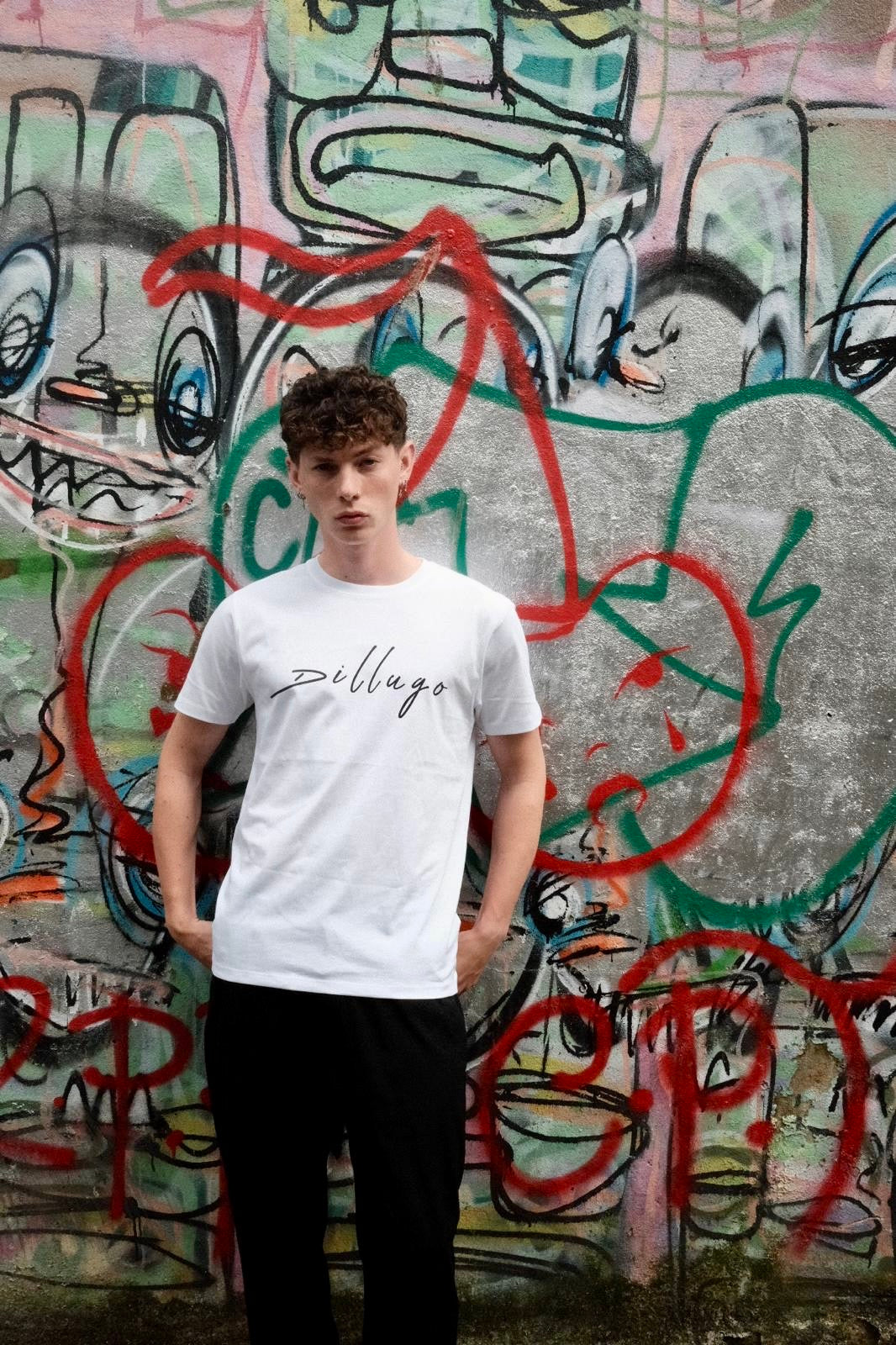 Male model wearing white t-shirt with "Dillugo" written across it (in black) on a graffiti backdrop.